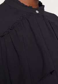 Blusa nera con un colletto alto e volant, dettagli decorativi con bottoni e un tessuto morbido e fluido. Presenta accentuazioni plissettate lungo la parte anteriore.