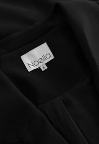 Črn material z gladko teksturo, s oznako, ki pravi "Noella" in velikostjo "S". Ovratnik je čist, brez vidnih okraskov.