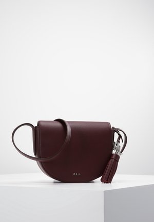 Bolso bandolera de cuero marrón oscuro con solapa redondeada, correa ajustable, herrajes plateados y un adorno de borla sobre un pedestal blanco.