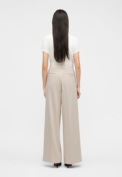 Γυναίκα που στεκεται πλάτη, με μακριά σκουρόχρωμα μαλλιά, φορώντας λευκό μπλουζάκι με κοντά μανίκια, μπεζ παντελόνι με φαρδιά μπατζάκια και ψηλοτάκουνα μαύρα παπούτσια.