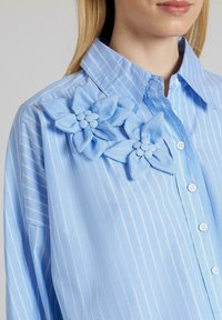 Camisa de botones a rayas de color azul claro que presenta dos grandes flores de tela texturizada en el hombro izquierdo, con un intrincado detalle en los pétalos.