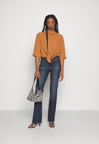 Camisa castanha de botões com a frente atada, jeans de boca de sino azul escuro, sandálias de salto alto pretas e uma bolsa com padrão de zebra. Tecido suave e texturizado.