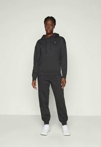 Frau steht mit schwarzem Puma-Hoodie und passender schwarzer Jogginghose sowie weißen Turnschuhen vor einem einfarbigen hellen Hintergrund.