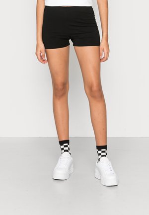 Person iført sort tætsiddende shorts, sort-hvide ternede sokker og hvide platformsneakers, der står på et gråt gulv.