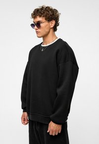 Zwarte oversized sweatshirt met een ronde halslijn, textuur stof en verlaagde schoudernaden, gecombineerd met zwarte broek en zonnebril.