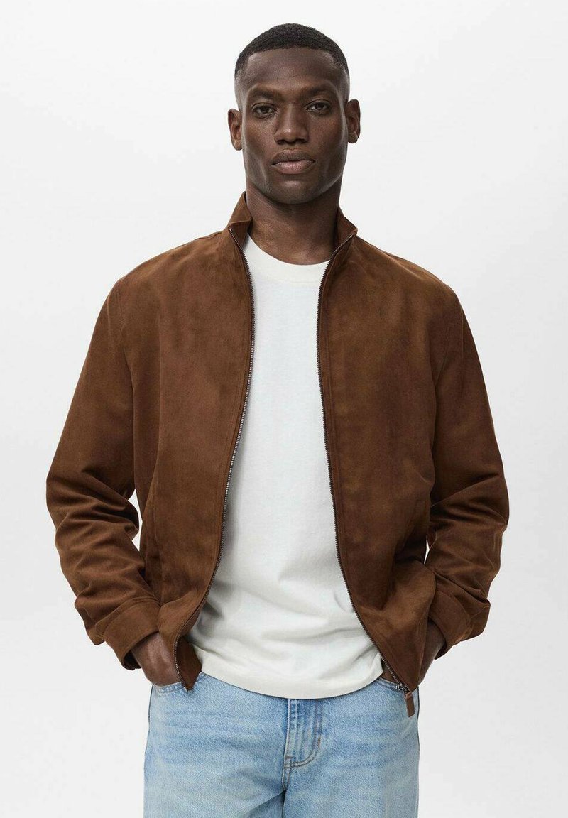 Chaqueta bomber de ante marrón con cuello alzado, cierre de cremallera en la parte delantera y bolsillos laterales, combinada con una camiseta blanca y jeans de color azul claro.