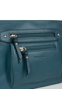 Borsa a mano in pelle color teal con numerose tasche con zip, accenti in metallo dorato e una superficie liscia. Presenta il logo del marchio impresso sulla parte anteriore.
