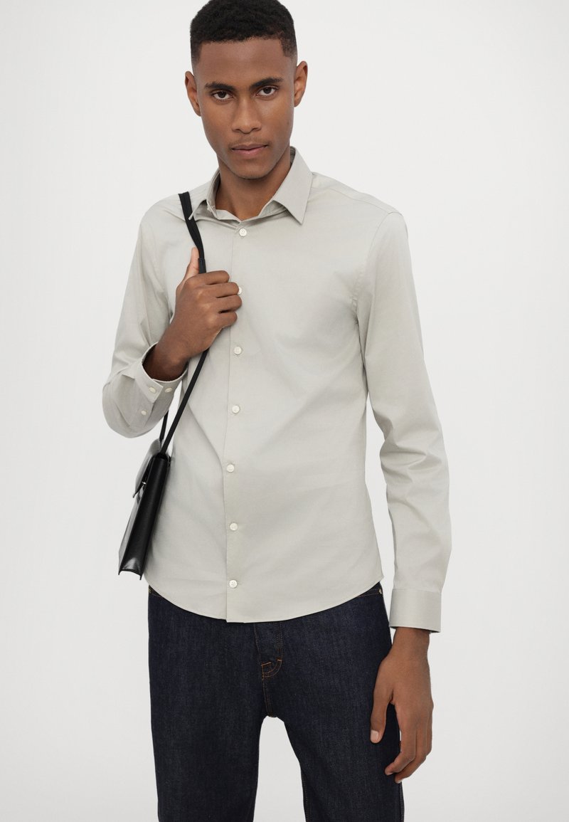 Chemise boutonnée gris clair avec col standard, manches longues et texture lisse, associée à un jean en denim bleu foncé.