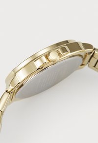 Goldfarbene Armbanduhr mit einem glatten Metallband, runder Gehäuseform und einem strukturierten Kronenbereich. Die Rückseite zeigt eingravierte Details mit einer flachen Oberfläche.