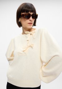 Sweatshirt de couleur crème avec des manches bouffantes, doté de deux petits nœuds au niveau du col et d'une finition texturée. Associé à des lunettes de soleil oversize en écaille de tortue.