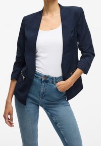 Blazer bleu marine à manches trois-quarts, design élégant, deux poches zippées, tissu léger, associé à un haut blanc et un jean bleu.