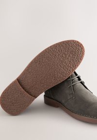 Dunkelgrauer Wildleder-Stiefelette mit Schnürdesign, brauner, strukturierter Gummisohle und kontrastierenden braunen Nähten entlang der Kante.