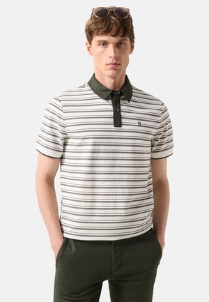 Unge mand iført en hvid og gråstribet polo med en mørkegrøn krave og mørkegrønne bukser, solbriller hvilende på hans hoved.