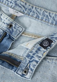 Jean denim bleu clair avec un motif délavé, braguette zippée, bouton argenté et accents cloutés. Coutures apparentes et texture douce.