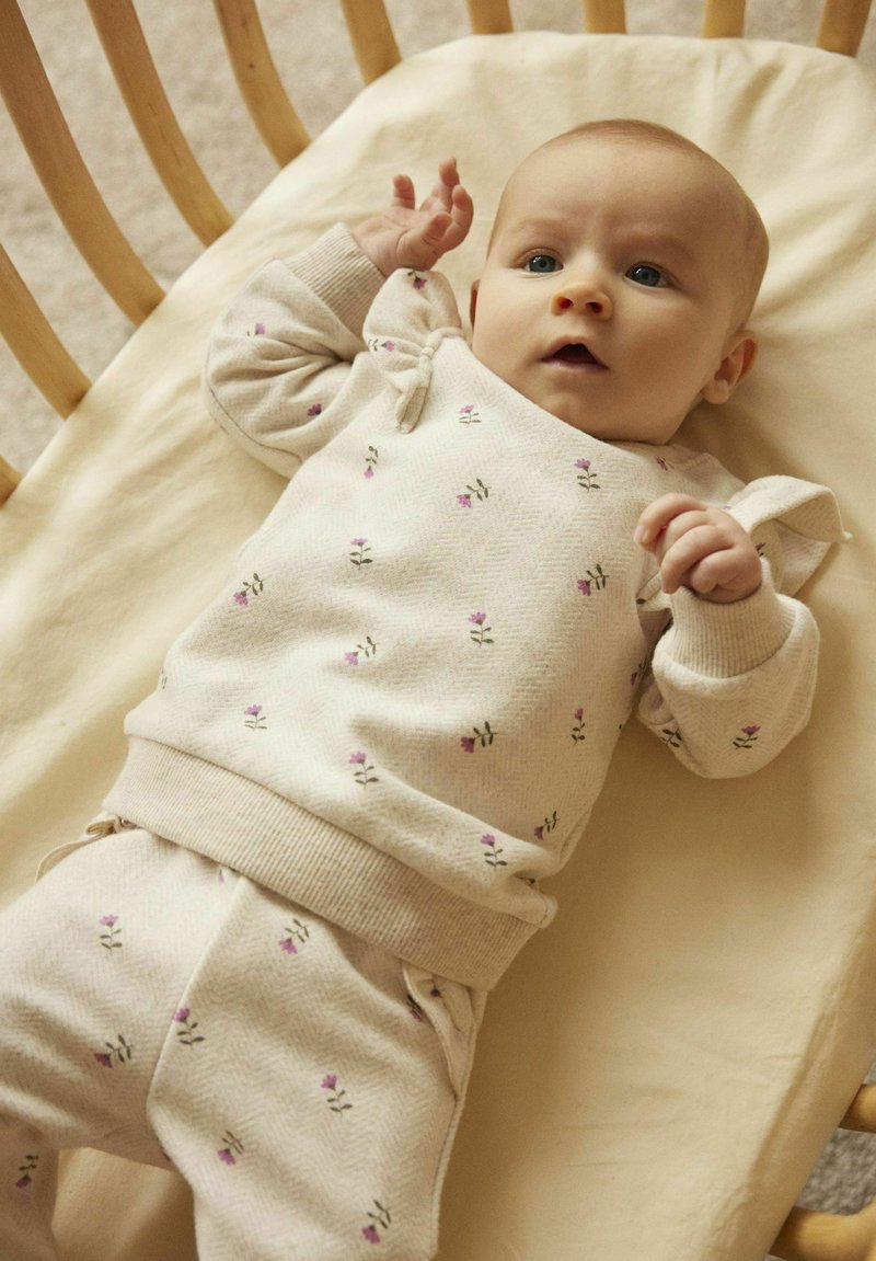 Completo per neonati di un beige chiaro, realizzato in tessuto a trama, con piccoli motivi floreali. La parte superiore ha polsini a costine e una vestibilità comoda, abbinata a pantaloni coordinati.
