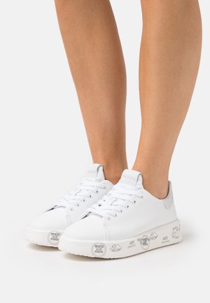 Weiße Leder-Sneaker mit niedrigem Schnitt, Schnürung vorne und Logoprints auf dicken Sohlen, getragen an den Füßen vor einfarbigem Hintergrund.