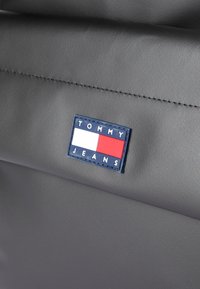 Pilkas tekstūruotas audinys su mėlynu stačiakampiu žymekliu, ant kurio užrašyta "TOMMY JEANS", raudonais ir baltais horizontaliais dryžiais bei matomais siūlės.