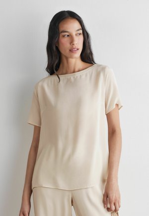Kvinde med langt mørkt hår iført en løs, kortærmet beige bluse og matchende bukser, stående op ad en ensfarvet hvid væg.