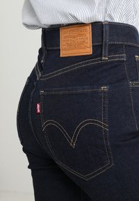 Jeans de denim azul escuro apresentam um patch de couro na parte de trás com marcação em relevo e costuras amarelas. Um design de bolso em forma de coração é visível.