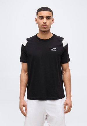 Άνδρας που φοράει μαύρο t-shirt EA7 με λευκές λεπτομέρειες στους ώμους και λευκό σορτς, στέκεται μπροστά σε απλό ανοιχτό γκρι φόντο.