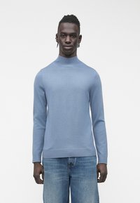 Svetlo modra pletena majica z visokim ovratnikom, dolgimi rokavi in rebrastimi manšetami ter spodnjim robom; kombinirana z modrimi kavbojkami iz jeansa.