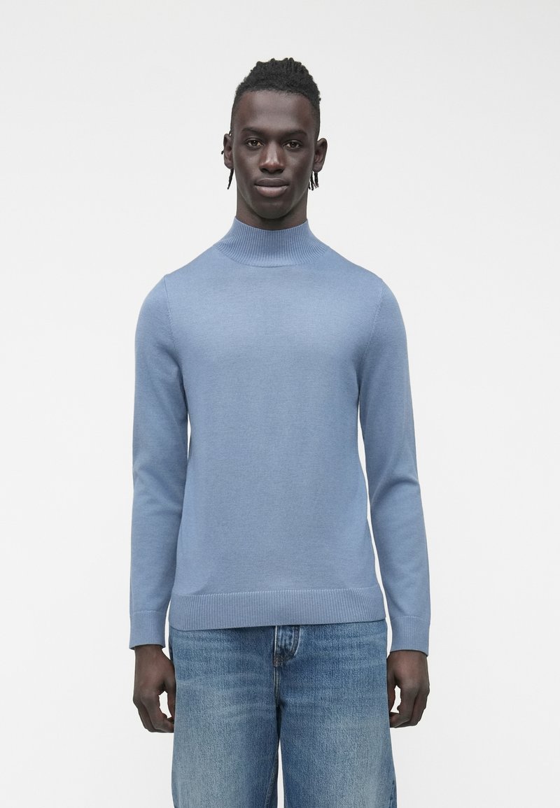 Svetlo modra pletena majica z visokim ovratnikom, dolgimi rokavi in rebrastimi manšetami ter spodnjim robom; kombinirana z modrimi kavbojkami iz jeansa.