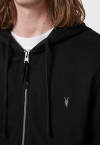 Black zip-up hoodie made of soft fabric, featuring a silver zipper and a small white goat skull embroidery on the left chest area.
