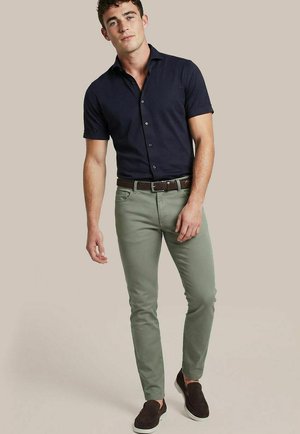 Junger Mann steht, trägt ein marineblaues kurzärmliges Hemd mit Knöpfen, hellgrüne Skinny-Hose, braunen Gürtel und dunkle Slip-On-Schuhe.