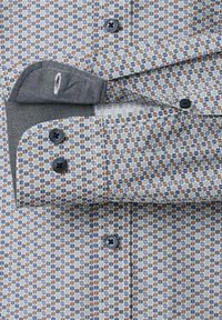 Close-up van een gestreepte manchetkraag met blauwe en bruine geometrische vormen en donkerblauwe knopen op lichte stof.