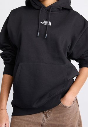 Osoba nosząca czarną bluzę z kapturem The North Face z logo na piersi, jedną ręką w kieszeni, w połączeniu z brązowymi spodniami, skróconymi na wysokości talii.