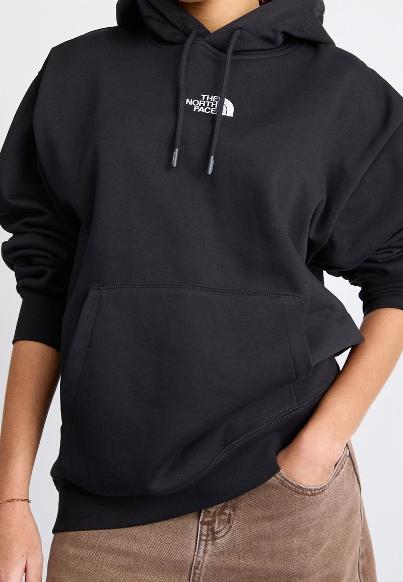 Personne portant un sweat à capuche noir The North Face avec un logo sur la poitrine, une main dans la poche, associé à un pantalon marron coupé à la taille.