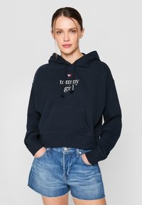 Felpa cropped blu navy con tasca frontale, cordino e testo "tommy girl". Abbinata a pantaloni corti di jeans azzurri.