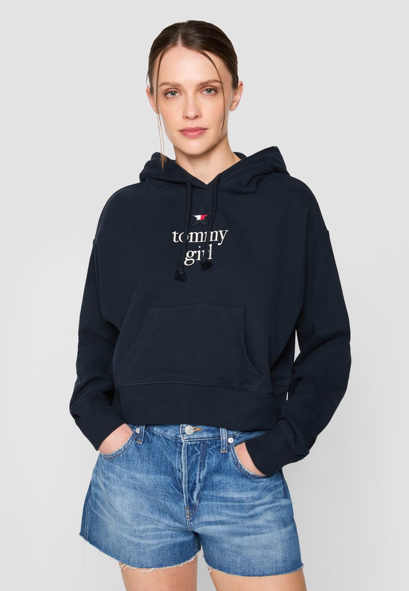 Felpa cropped blu navy con tasca frontale, cordino e testo "tommy girl". Abbinata a pantaloni corti di jeans azzurri.