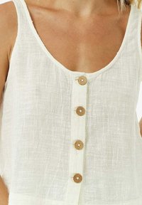 Débardeur en lin blanc avec un col rond plongeant et cinq boutons en bois sur le devant. Le tissu présente une texture tissée apparente.