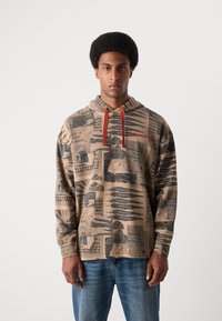 Hooded sweatshirt in beige with black and red graphic prints, including text and patterns. Features orange drawstrings and relaxed fit.
