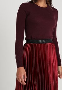 Bordeaux lange mouwen trui gecombineerd met een rode plooienrok met een gestructureerde zwarte tailleband. Soepele brei- en glanzende stofdetails.