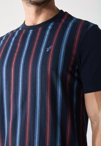 Tumedat sinise lühikeste varrukatega T-särk, millel on vahelduvad punased ja sinised vertikaalsed triibud ning rinnal väike tikanditega logo.