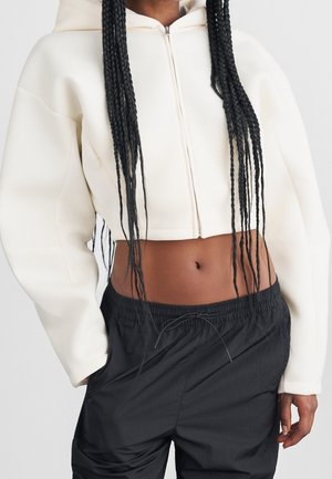 Osoba z długimi splecionymi włosami, ubrana w krótki biały zip hoodie i czarne spodnie z elastycznym pasem, ręce w kieszeniach, na jednolitym jasnym tle.