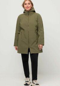 Manteau softshell vert olive avec capuche, fermeture éclair sur le devant, deux poches latérales et design mi-long. Porté sur un pantalon noir et des baskets.