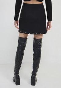 Donna che indossa una mini gonna nera con occhielli metallici all'orlo e stivali alti neri con tacco in pelle, in piedi su uno sfondo neutro.