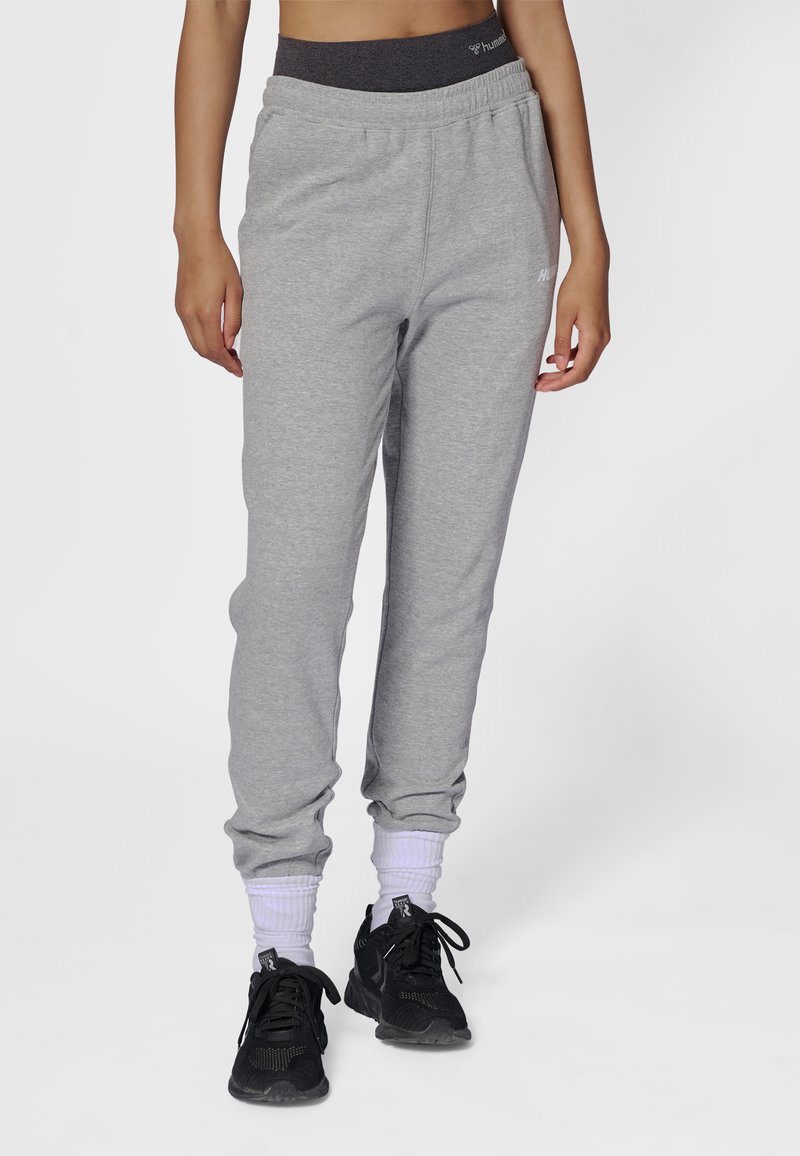 Grå sweatpants i bomuldsblanding med elastisk talje og manchetter, med logo på siden. Kombineret med sorte sportslige sko og hvide sokker.