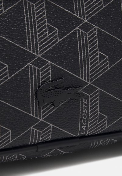 Un matériau noir texturé présentant un motif géométrique en gris. Un logo Lacoste noir est positionné de manière proéminente dans le design.