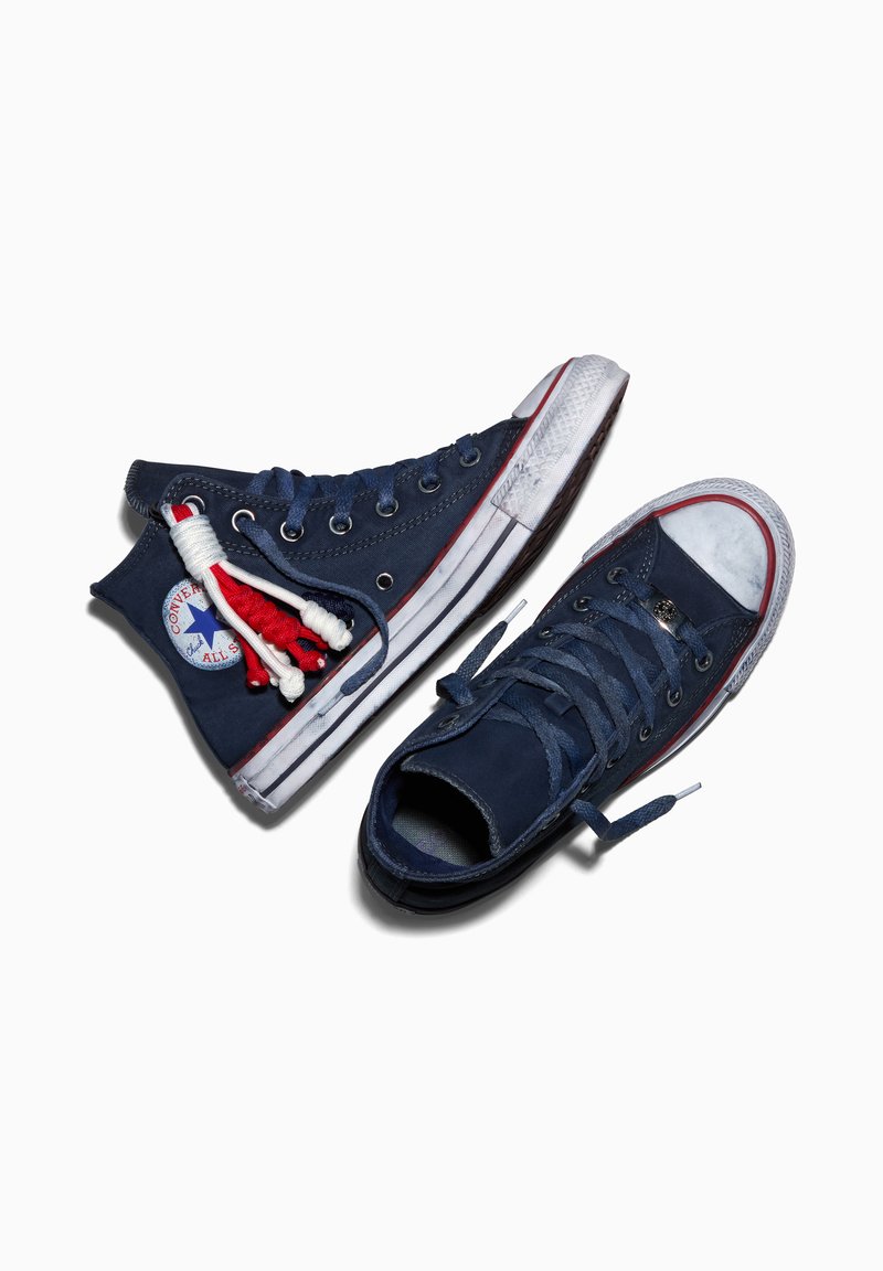 Paar dunkelblaue High-Top Converse-Sneaker, einer mit rot-weißen geflochtenen Schnürsenkeln, weiße Gummisohle mit rotem Streifen, auf weißem Hintergrund.