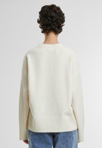Cream-colored oversized sweater with a ribbed hem and round neckline, made from textured, soft material. Casual fit with dropped shoulders.