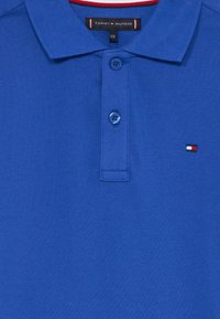 Μπλε πόλο μπλούza Tommy Hilfiger με δύο μπροστινά κουμπιά, γιακά και μικρό λογότυπο σε κόκκινο, άσπρο και μπλε χρώμα στην αριστερή πλευρά του στήθους.