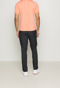 Man i korallfärgad kortärmad skjorta, svarta byxor och vita sneakers som står mot en enkel ljus bakgrund, vänd bort.