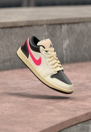 Zapatilla con colores beige, negro y rosa, con un logo Nike rosa y un logo Jordan negro, flotando sobre una superficie de hormigón.