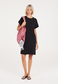 Schwarzes kurzärmeliges Kleid mit lockerer Passform, kombiniert mit einer pinken Tragetasche, die ein gestreiftes Handtuch hält. Das Model trägt schwarze Flip-Flops.