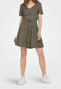Kurzes Kleid in olivgrün mit Leopardenmuster, verfügt über einen V-Ausschnitt, eine taillierte Taille und einen ausgestellten Rock, kombiniert mit weißen Sneakers.