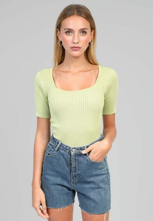 Femme portant un haut à manches courtes côtelé vert clair avec un décolleté carré et un short en denim bleu taille haute, debout devant un fond gris uni.